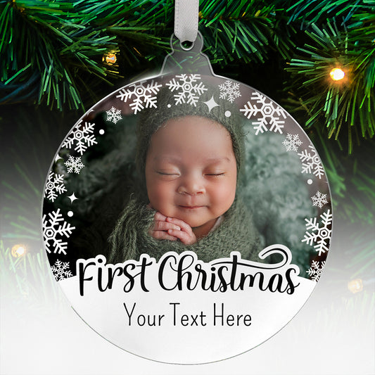 Round acrylic Christmas tree ornament with a photo uloaded of a baby and decorative white snowflakes around the border, featuring the words 'First Christmas' and below 'Your Text Here' in a tall font, hanging on a tree with green branches and fairy lights.