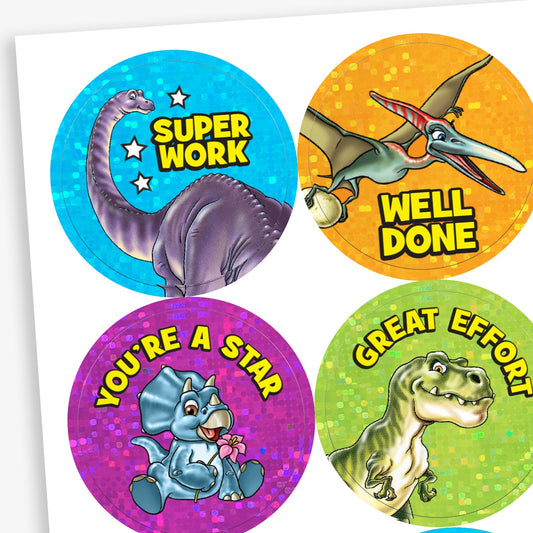 Sheet of holographic circular reward stickers for children, each featuring a cartoon dinosaur and motivational text. Includes:a long-necked dinosaur with the words 'SUPER WORK' on a blue sparkly background; a flying pterodactyl with 'WELL DONE' on an orange background: a smiling baby triceratops holding flowers, with the text 'YOU'RE A STAR' on a purple background; and a green T. rex with the phrase 'GREAT EFFORT' on a yellow-green sparkly background