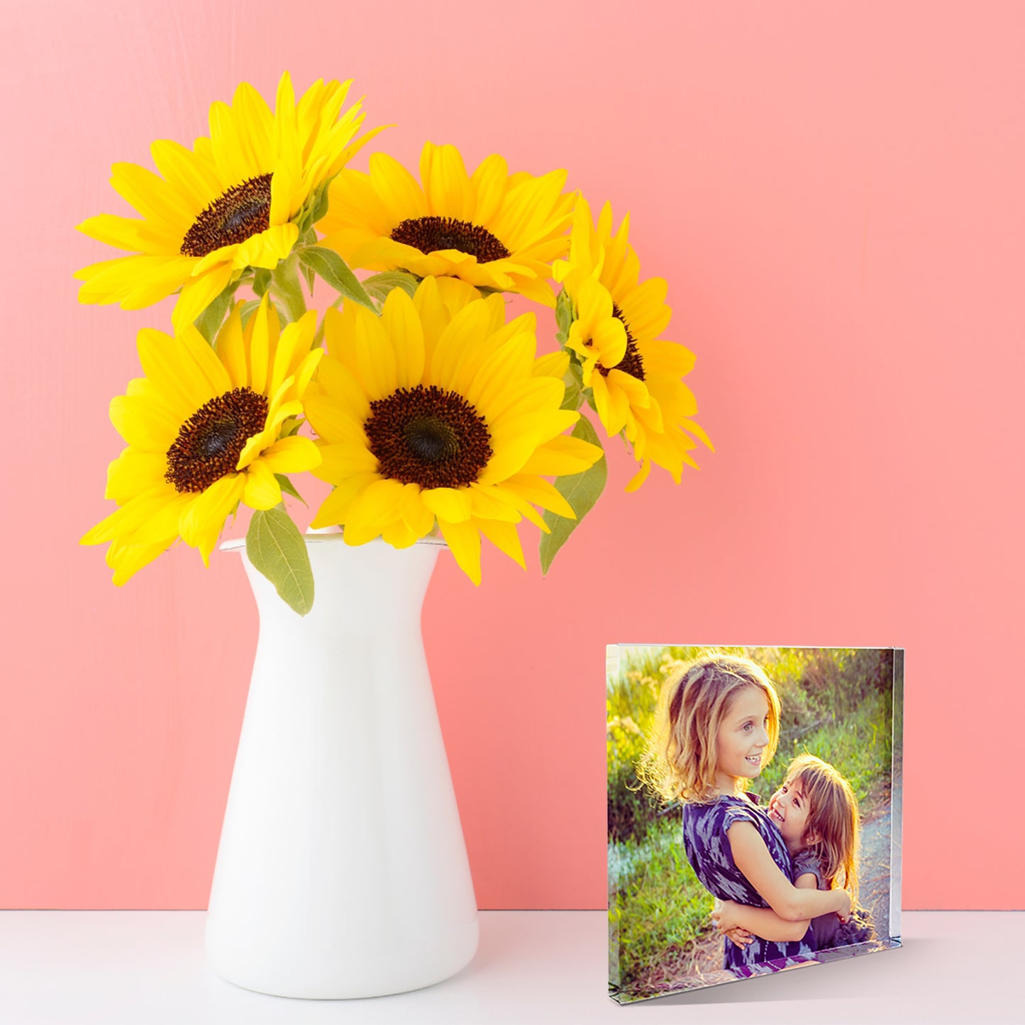 A square acrylic photo block featuring a family photo. The image shows the full areas printed with the photo of two smiling children. The block has a glossy, freestanding design ideal for display. The photo bloack is stood next to a vase of sunflowers.