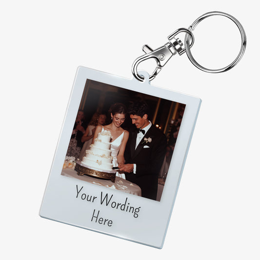 A personalised keyring featuring a rectangular photo frame design with a metal keychain and clasp. The photo shows a smiling bride and groom cutting their wedding cake. Below the image, there is placeholder text reading 'Your Wording Here'. The keyring has a glossy finish and a polaroid-style layout.