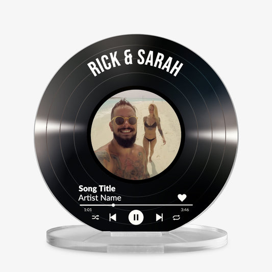 Acrylic custom vinyl record-style display with a circular photo in the centre, personalised text 'Rick & Sarah' at the top, and a mock music player interface showing song title and artist name at the bottom.