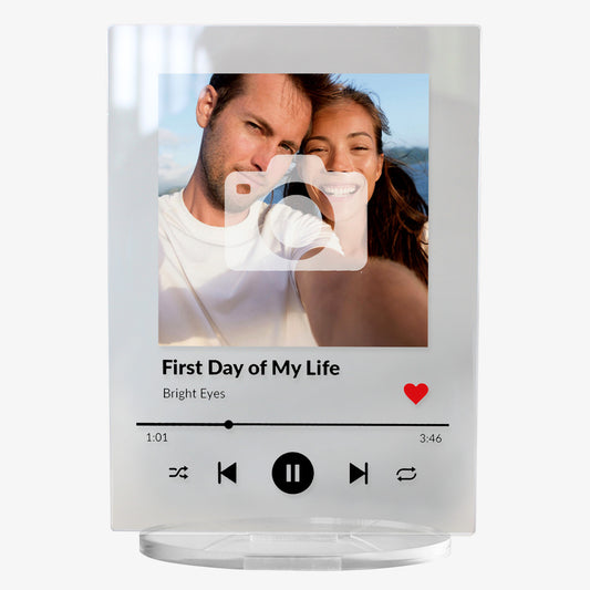 A personalised acrylic music plaque styled like a digital music player. It features a photo of a smiling couple at the top, followed by song details: 'First Day of My Life' by Bright Eyes. Music player controls, a progress bar, and a red heart icon appear below, mimicking a streaming app interface. The plaque stands upright on a clear circular base.