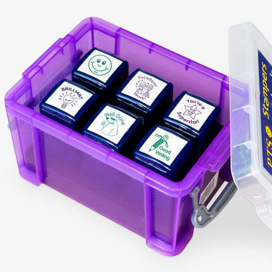A transparent purple storage box containing six teacher stampers with positive feedback phrases and images. The stamps read: 'BRILLIANT!' with a light bulb, 'Well Done' with a smiling face, 'Good Work' with a pencil, 'Excellent Work' with a star character, 'You're a Superstar!' with a starburst, and a smiley face icon. The clear lid is slightly open, displaying the 'PTS Stampers' label.