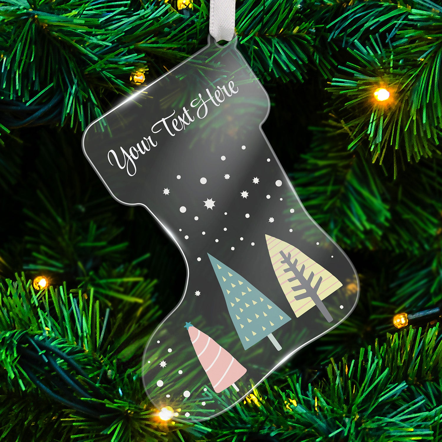 Clear acrylic Christmas stocking-shaped tree ornament decorated with a snow scene falling on 3 christmas trees in pale  colours of pink, green and yellow, featuring a space at the top for a customisation saying 'Your Text Here' in white script, hanging on a Christmas tree.