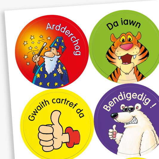 Sheet of circular stickers featuring cartoon characters and Welsh praise phrases. Includes: wizard with stars on a red background and the word 'Ardderchog'; A smiling tiger on a green background with the phrase 'Da iawn';  A thumbs-up hand on a yellow background with the phrase 'Gwaith cartref da'; A polar bear giving a thumbs up on a purple background with the word 'Bendigedig!'