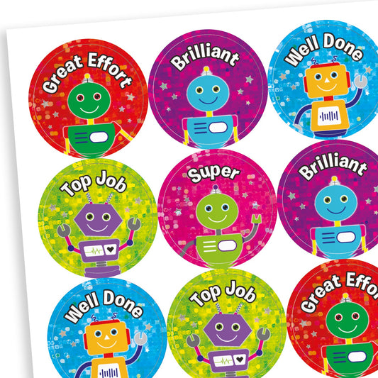 A sheet of colourful, holographic reward stickers featuring cheerful cartoon robots. Each circular sticker displays a positive message such as 'Great Effort', 'Brilliant', 'Well Done', 'Top Job', and 'Super'. The backgrounds are bright and sparkly in colours like red, purple, blue, green, and pink, with each robot in a contrasting colour for visual appeal.