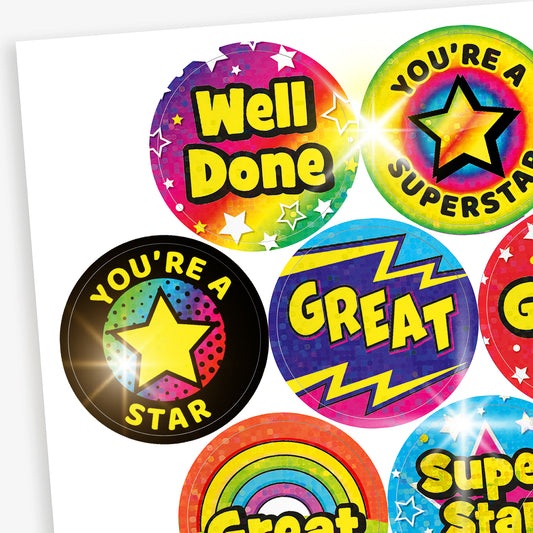 A selection of colourful circular reward stickers with messages such as 'Well Done', 'You're a Superstar', 'You're a Star', 'Great', and 'Super Star'. The stickers feature bold, playful fonts and vibrant holographic backgrounds with stars, lightning bolts, and rainbows.