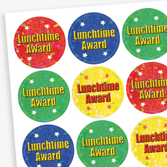 A sheet of 25mm round holographic 'Lunchtime Award' stickers. Each sticker features a vibrant, reflective background in colours such as red, blue, green, and yellow, with the text 'Lunchtime Award' in bold red or yellow letters. Ideal for recognising positive behaviour during lunchtime.