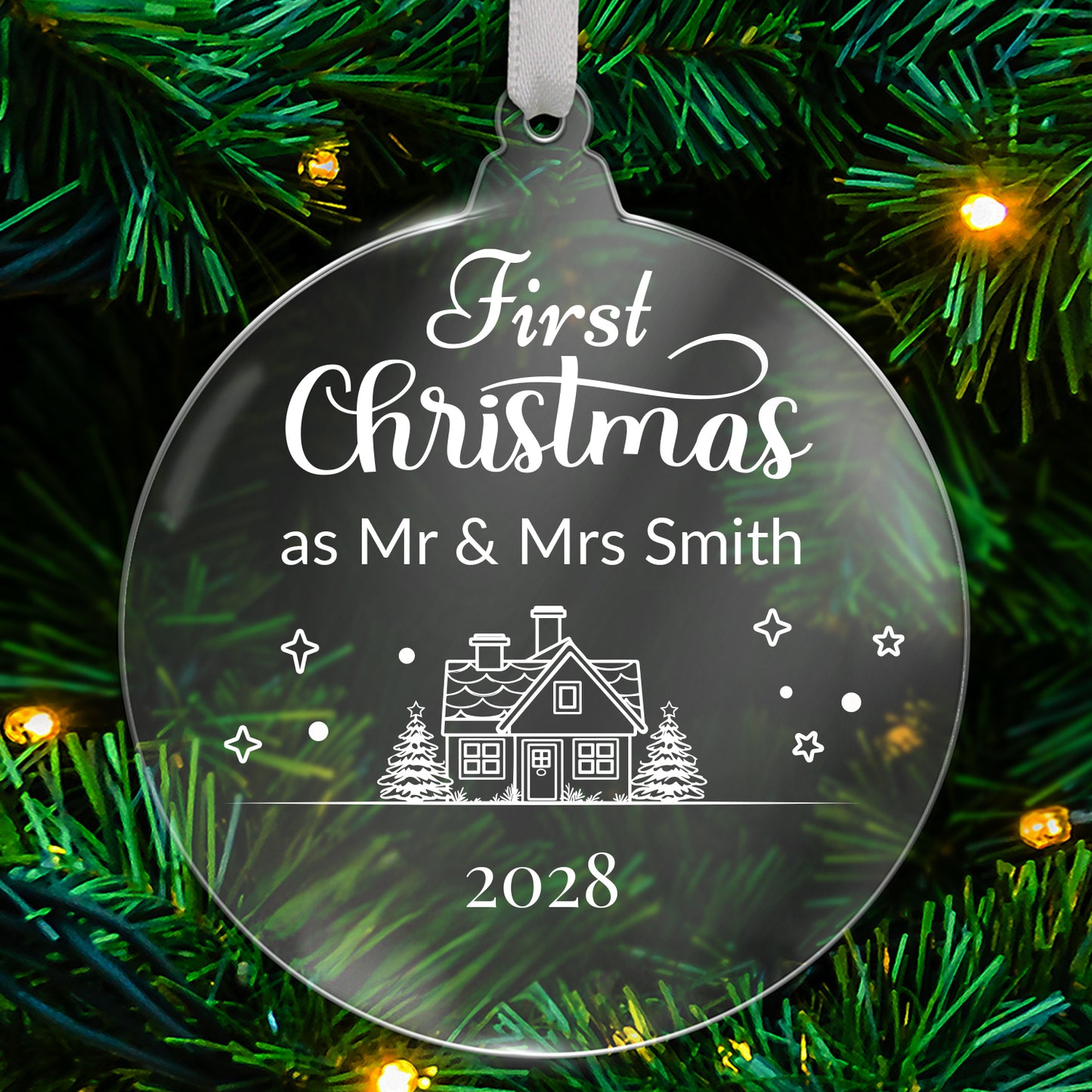 Round clear acrylic Christmas tree ornament with white design reading 'First Christmas as Mr & Mrs Smith 2028', featuring a house with trees, stars, and snowflake details, hanging on a green tree with fairy lights.