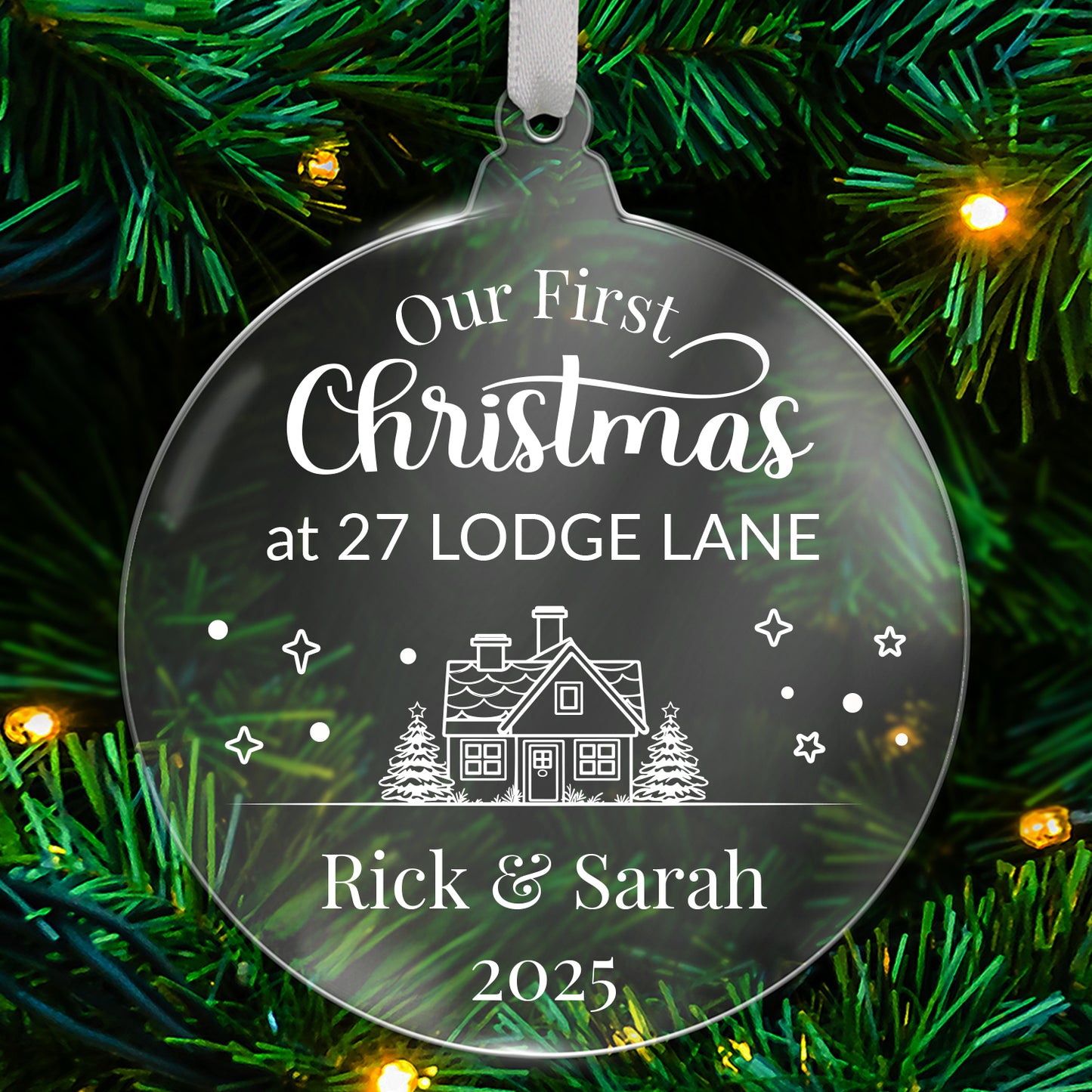 Round clear acrylic Christmas tree ornament with white design reading 'Our First Christmas at 27 LODGE LANE', featuring a house with trees, stars, and snowflake details, with bottom customisable text 'Rick & Sarah 2025' hanging on a green tree with fairy lights.