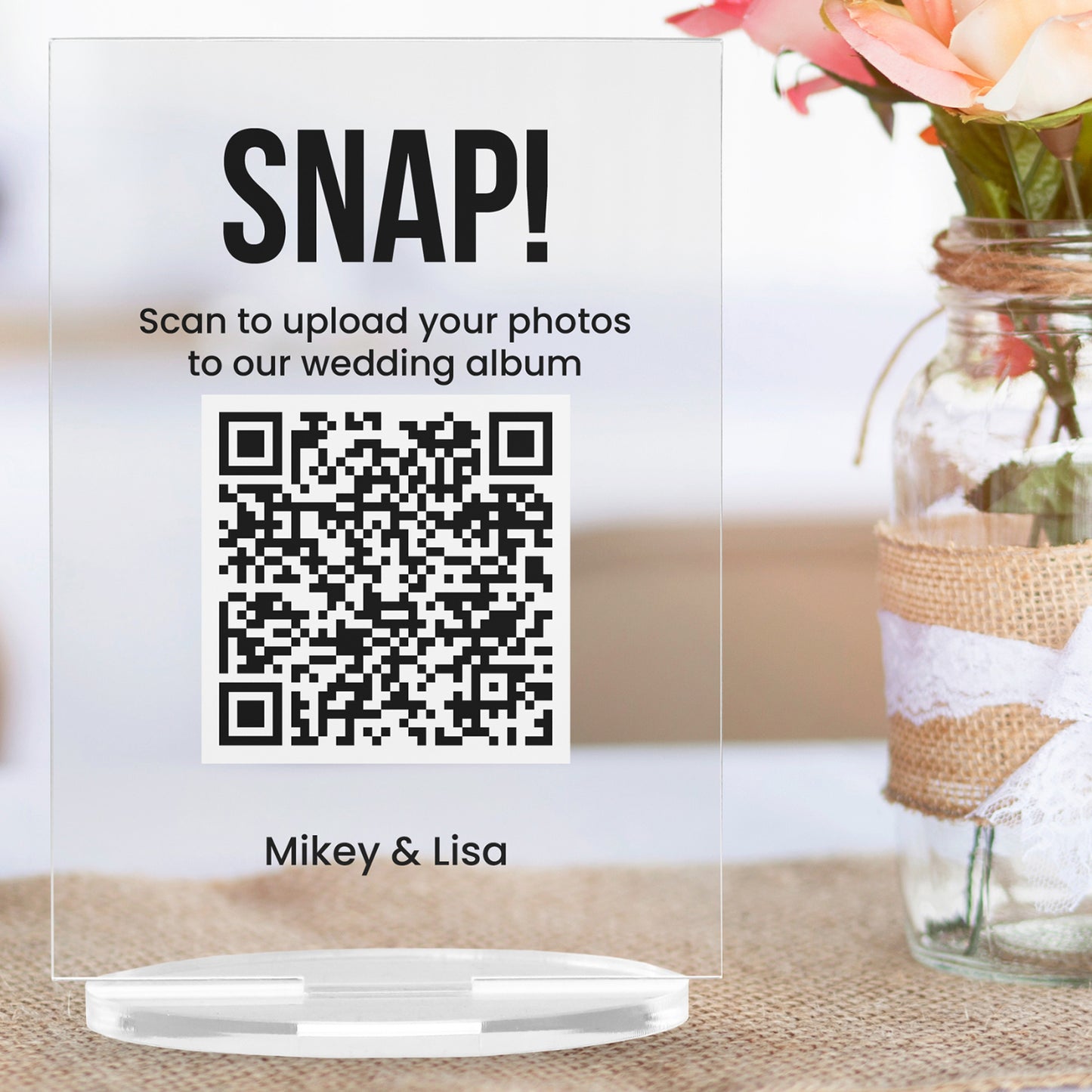 A clear acrylic sign with bold black text at the top reading 'SNAP'. Below, it another placeholder 'Scan to upload your photos to our wedding album'. A large black and white QR code is centrally displayed, and the placeholder 'Mikey & Lisa' appears at the bottom. The sign stands upright on a circular clear base.