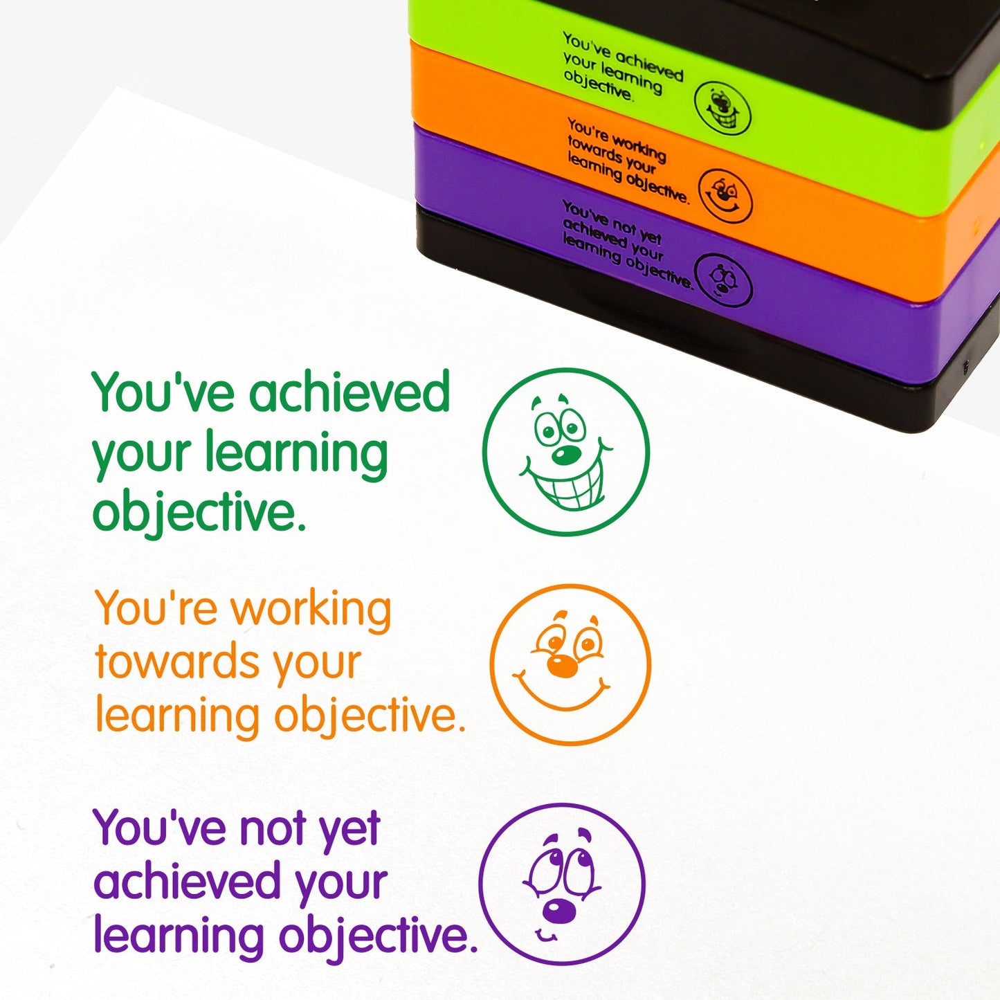 3 stacked rectangular self-inking stampers measuring 44mm by 13mm, featuring the imprint - 'You've achieved your learning objective' across 3 lines in green ink with a beaming emoji face to the right; 'You've not yet achieved your learning objective' across 3 lines in purple ink with a sorry emoji face to the right; 'You're working towards your learning objective' across 3 lines in orange ink with a smiling emoji face to the right