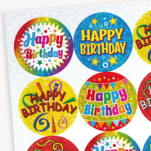 A sheet of colourful, holographic circular 'Happy Birthday' stickers with celebratory designs. Each sticker features bright backgrounds in blue, yellow, and red, decorated with confetti, balloons, and streamers, with bold 'Happy Birthday' text in various styles.