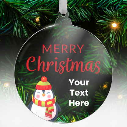 Round acrylic Christmas tree ornament featuring a festive penguin wearing a hat and scarf, with the message 'Merry Christmas' in red and space for custom text below, hanging on a tree with green needles and lights.