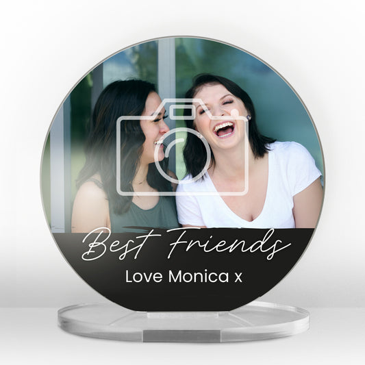 A round acrylic display piece with a photo of two friends sharing a moment. A placeholder camera icon is overlaid on the image, showing a customisable photo area. The bottom section has a black band with the words 'Best Friends' in cursive and space for personalised text reading 'Love Monica x. The item is displayed on a clear round stand.