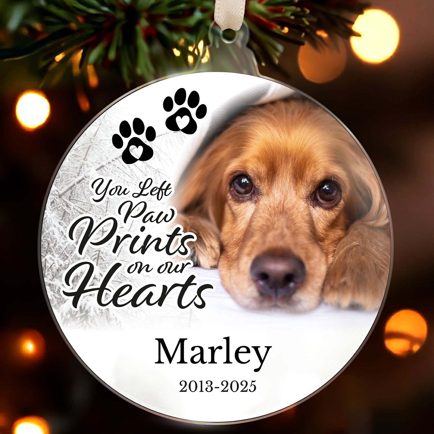 Memorial ornament with a dog's face and text 'You Left Paw Prints on our Hearts' and 'Marley 2013-2025' against a blurred Christmas tree background.