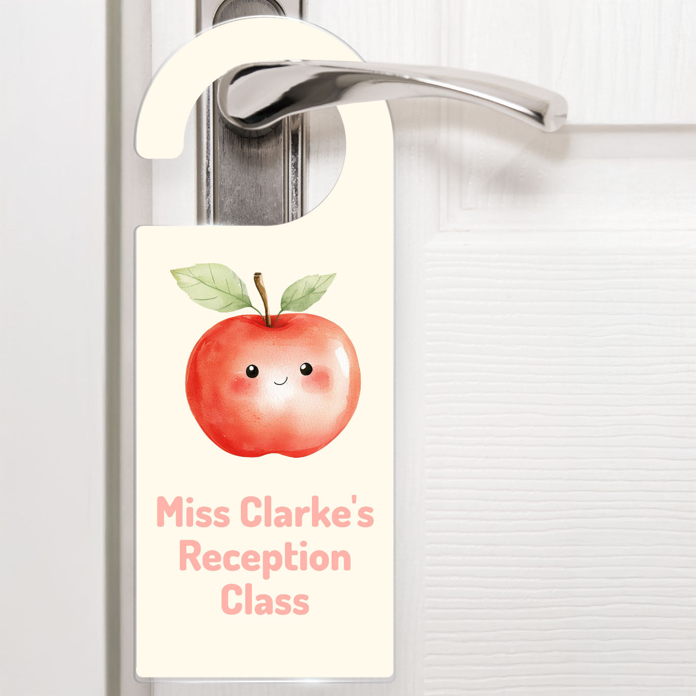 A cream door hanger hanging on a silver handle, featuring a cute watercolour apple above the text 'Miss Clarke's Reception Class' in soft pink letters. The design is playful and child-friendly, ideal for decorating a teacher's classroom door.