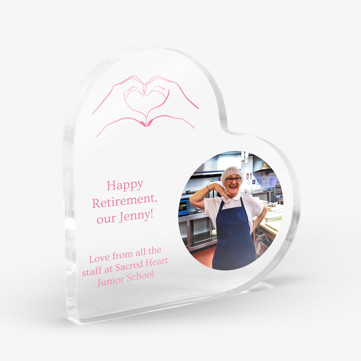 Heart-shaped acrylic block with a photo of a person in a kitchen, an image of hands making a heart shape and text 'Happy Retirement our Jenny' 'Love from all the staff at Sacred Heart Junior School'. Block is shown on a white background.