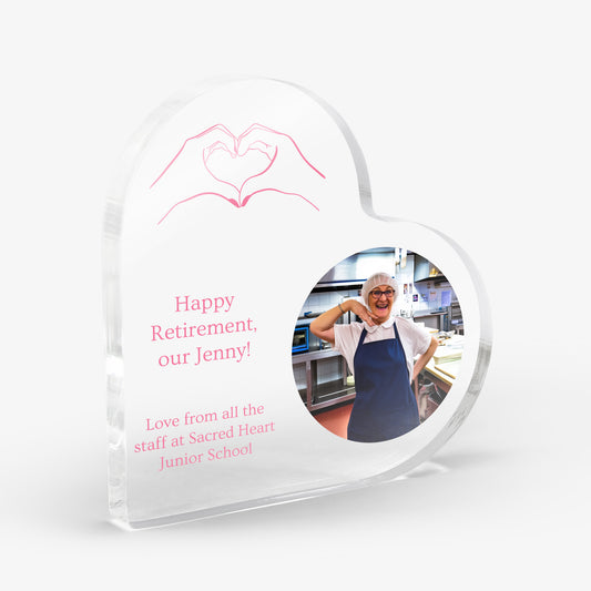 Heart-shaped acrylic block with a photo of a person in a kitchen, an image of hands making a heart shape and text 'Happy Retirement our Jenny' 'Love from all the staff at Sacred Heart Junior School'. Block is shown on a white background.