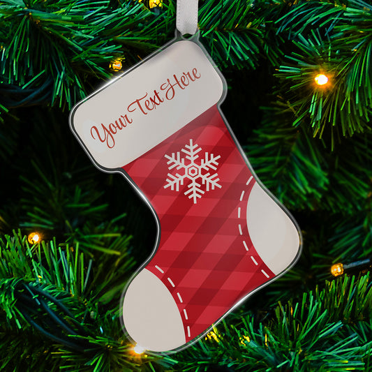 Personalised acrylic Christmas stocking-shaped tree ornament decorated with a red tarten style background with a white snowflake on it, and white top, heel and toe of the stocking. Featuring a space at the top for a customisation saying 'Your Text Here' in red script, hanging on a Christmas tree with lights.