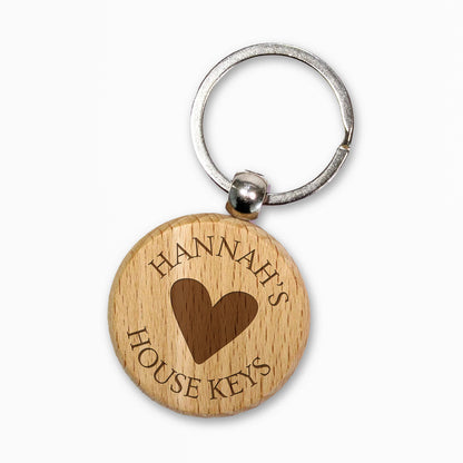 Circular wooden keyring with a metal ring, engraved with text for customisation saying 'Hannah's' a central image of a heart etched and underneath 'House Keys' 